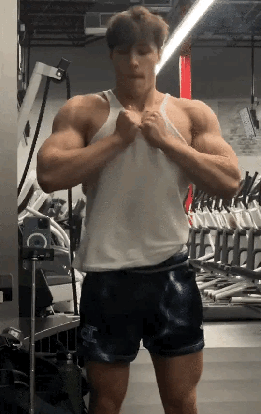 A muscular man in a tank top and shorts flexes in a gym.