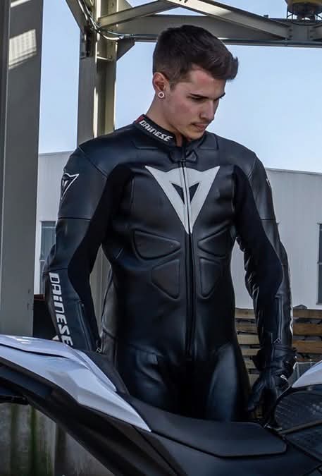 A young man wearing a full black and white Dainese leather motorcycle suit and matching gloves stands outdoors next to a bike.