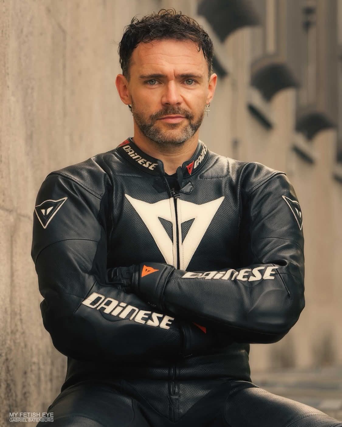 A mature bearded man in a full black and white Dainese leather motorcycle suit poses outdoors with his arms crossed.