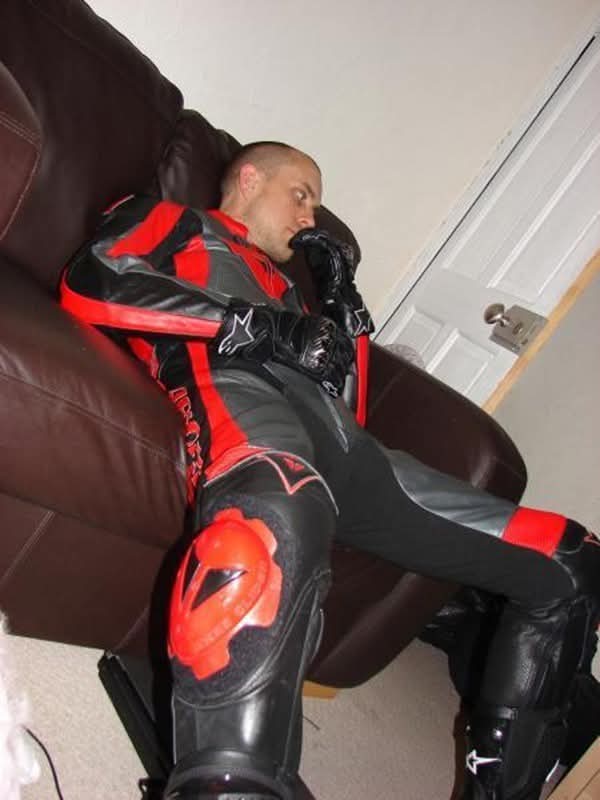 A man with a buzzcut reclines on a leather chair while wearing a full black and red leather motorcycle suit, gloves, and boots.