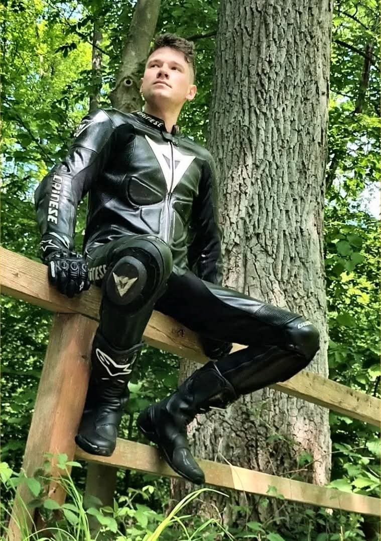 A young man in a full Dainese leather motorcycle suit, gloves, and boots sits on a wooden fence in a wooded area.